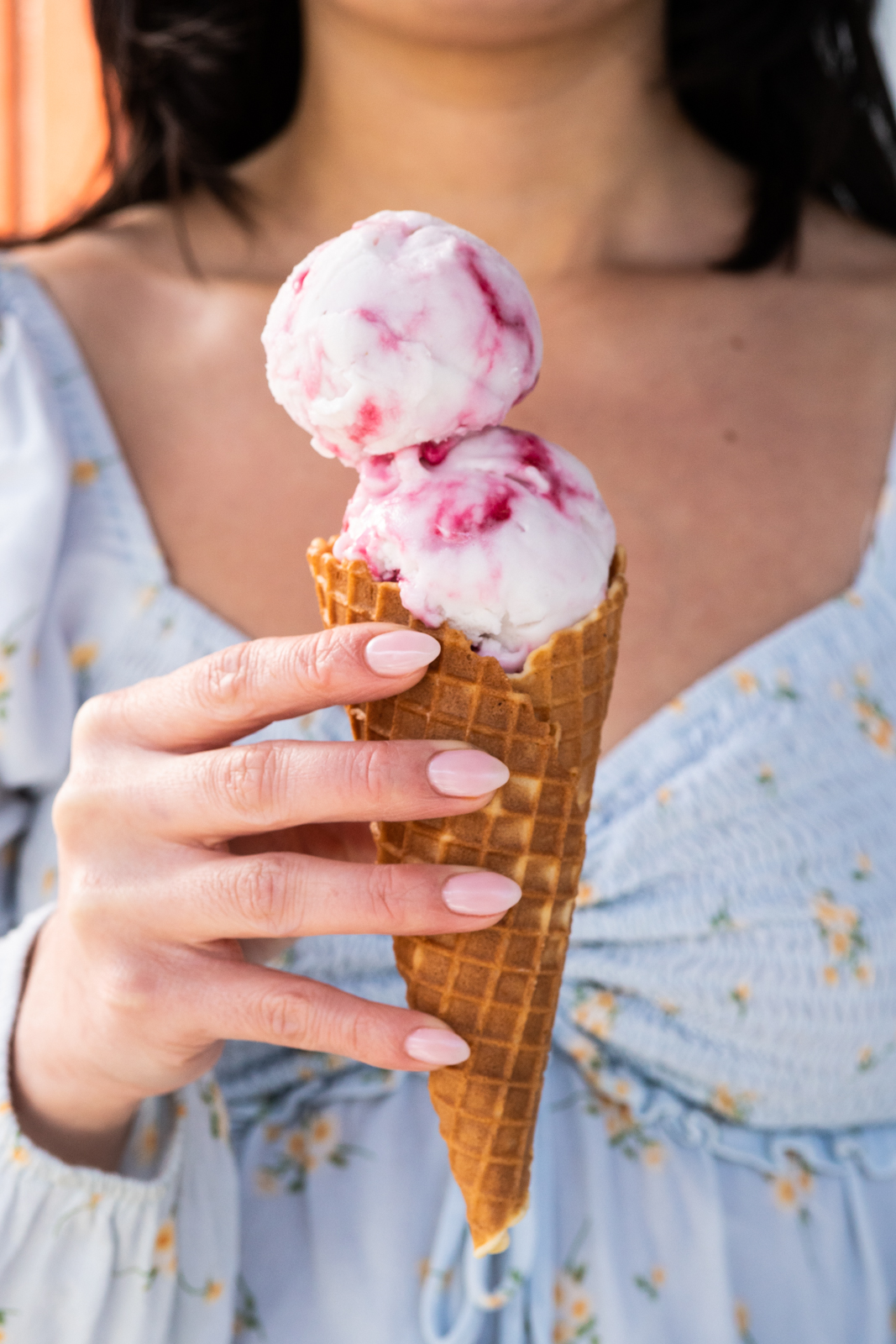Woman's hand holding a vanilla cone topped with 2 scoops of Dairy-Free Coconut Hibiscus ice cream