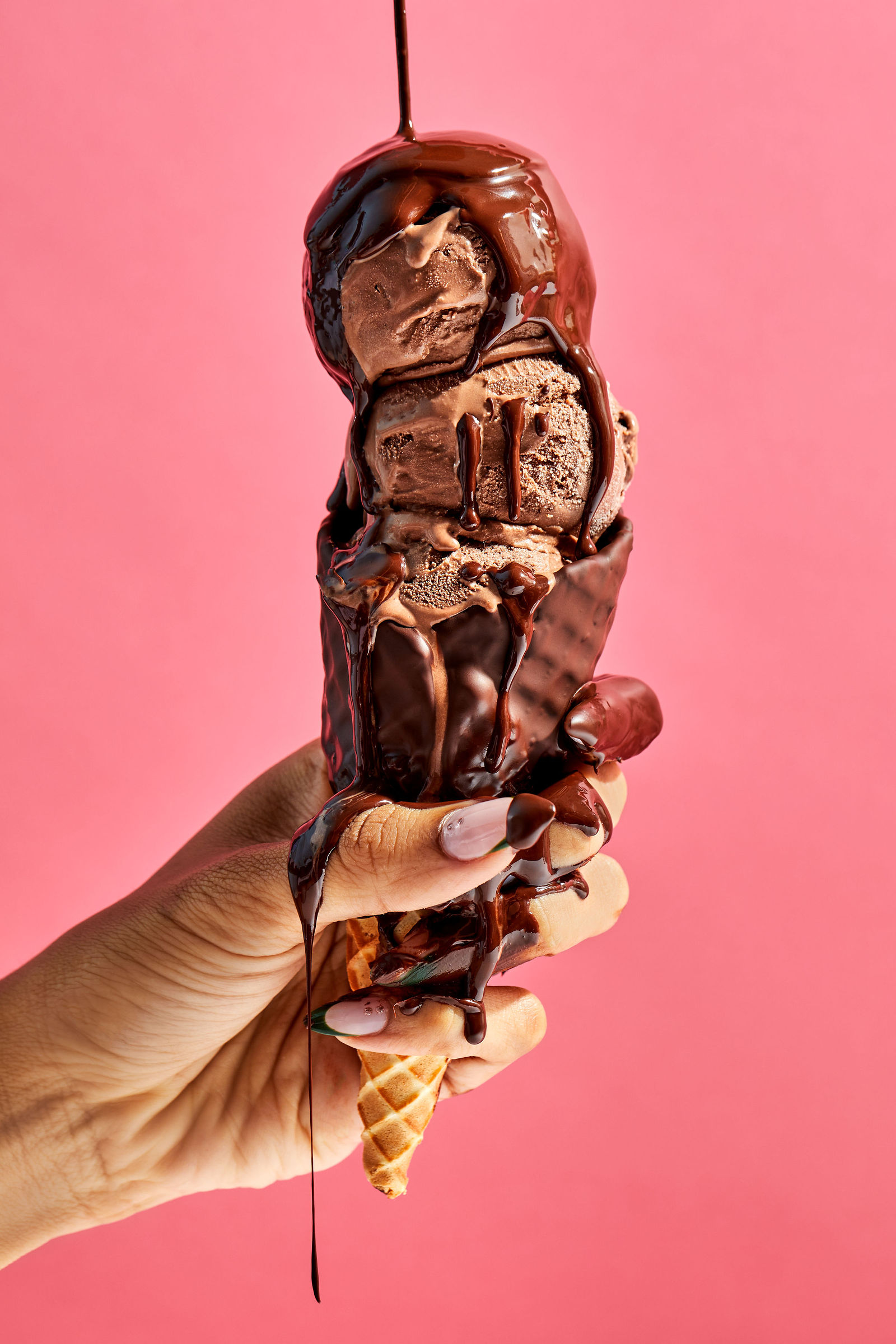 hands holding a chocolate cone with hot fudge dripping down scoops of chocolate ice cream